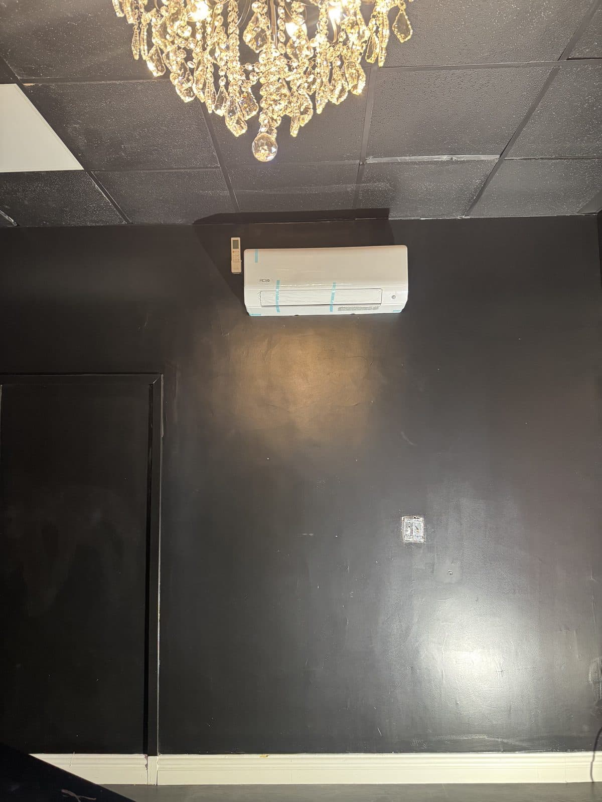 Mini-split indoor unit on a black wall beneath a crystal chandelier, beside a dark door