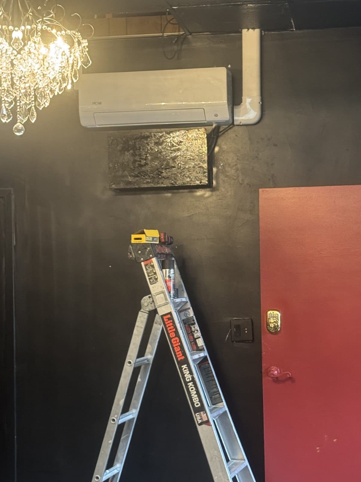 Mini-split indoor unit beside a red door with line-set cover running to the ceiling, ladder in front