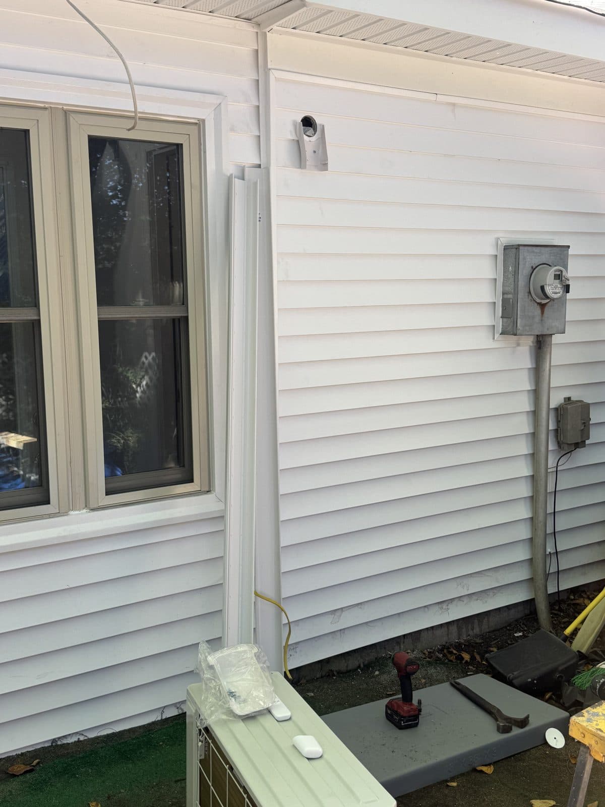 Exterior wall with a white line-set cover running down the siding from the sealed wall sleeve to the outdoor unit on its pad